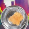 Golden cannabis concentrate in a clear jar against a colorful cityscape background.